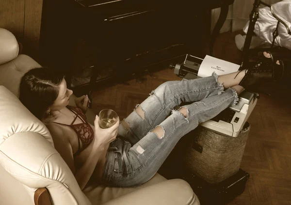 Jonge mooie vrouwelijke schrijver in rode beha en jeans zittend op een bank zet haar benen op retro typemachine — Stockfoto