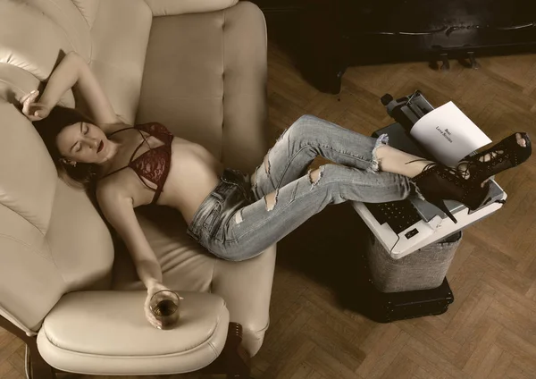 Молодая красивая писательница в красном бюстгальтере и джинсах, сидящая на диване, положила ноги на пишущую машинку — стоковое фото