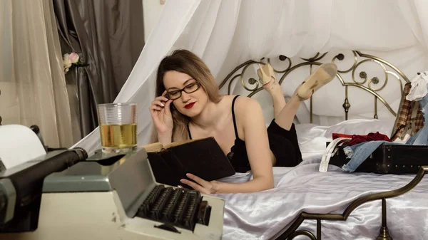 Svůdně mladá žena ležící na posteli a čte si knihu. krásná ženská spisovatelka odpočívá, v popředí je retro psací stroj — Stock fotografie