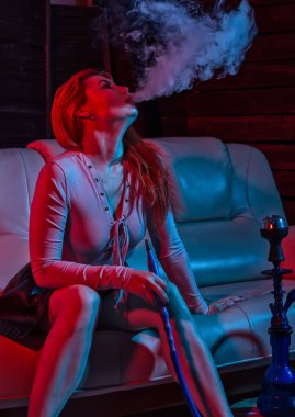 Seksi kızıl saçlı kadın kırmızı mavi tonları elektronik nargile vaping. kız bir kanepede oturan ve darbeler beyaz doom