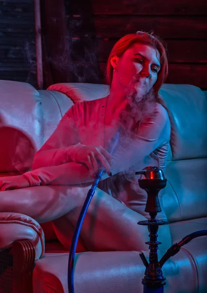 Mulher ruiva sexy vaping cachimbo eletrônico em tons azuis vermelhos. menina sentado em um sofá e golpes branco desgraça — Fotografia de Stock
