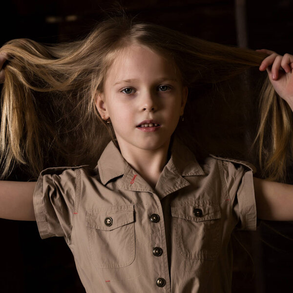 funny lovely little blonde child with different emotions on a dark background. beautiful schoolgirl holding herself hair