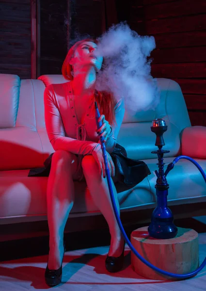 Seductora pelirroja sentada en un sofá y fuma narguile en tonos azules rojos —  Fotos de Stock