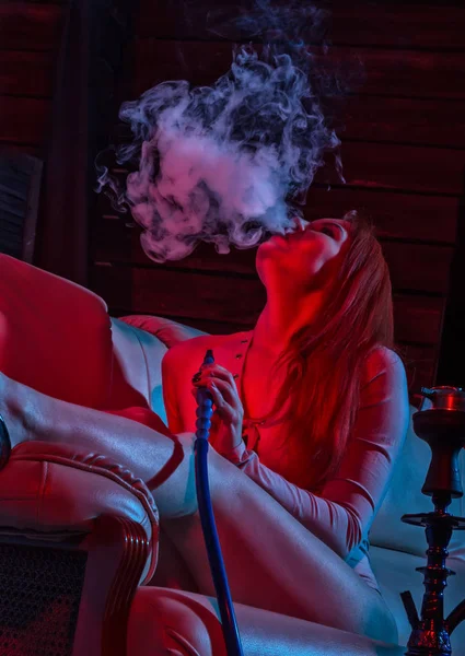 Csábító vöröshajú nő ül a kanapén, és dohányzik vízipipa piros kék árnyalatai — Stock Fotó