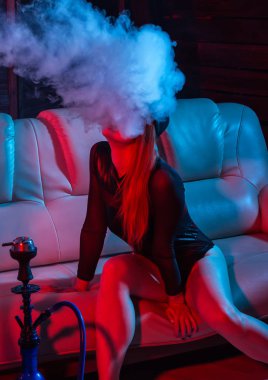siyah bodysuit seksi kızıl saçlı kadın bir kanepede oturan ve nargile sigara, kız büyük duman, kırmızı mavi tonları dışarı darbeler