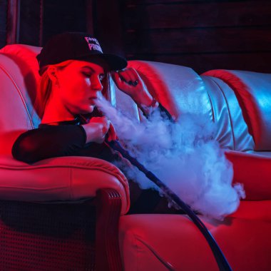 siyah bodysuit seksi kızıl saçlı kadın bir kanepede oturan ve nargile sigara, kız büyük duman, kırmızı mavi tonları dışarı darbeler