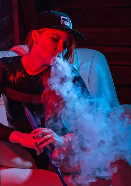 Sexy redhead woman in black bodysuit sitting on a sofa and smokes hookah, girl blows out big smoke, red blue tones — Stock Photo, Image