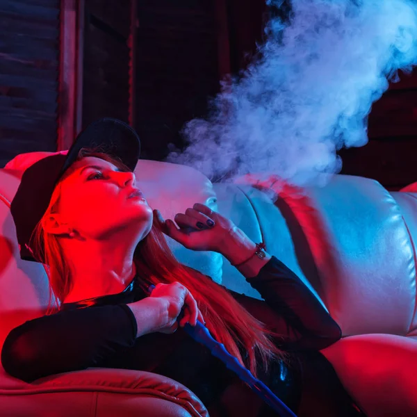 Sexy ruiva mulher no preto bodysuit sentado em um sofá e fuma narguilé, menina sopra para fora grande fumaça, tons azuis vermelhos — Fotografia de Stock