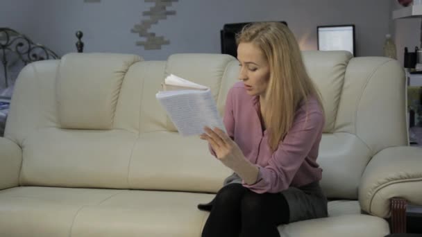 Blondine auf dem Sofa liest Notizen in Notizbuch und checkt Notizen mit dem Smartphone — Stockvideo