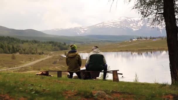 Backview av två vuxna killar, sitts på bänken och tillbringa tid genom att titta runt på skönheten i Mountain Lake — Stockvideo