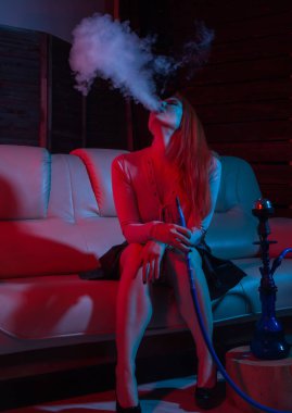 baştan çıkarıcı kızıl saçlı kadın kanepede oturan ve kırmızı mavi tonları nargile içiyor