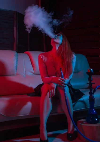 Sedutor ruiva mulher sentada em um sofá e fuma narguilé em tons azuis vermelhos — Fotografia de Stock