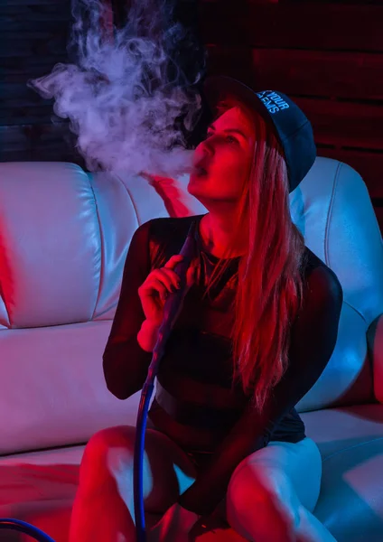 Sexy rossa donna in body nero seduto su un divano e fuma narghilè, ragazza soffia fuori grande fumo, rosso toni blu — Foto Stock