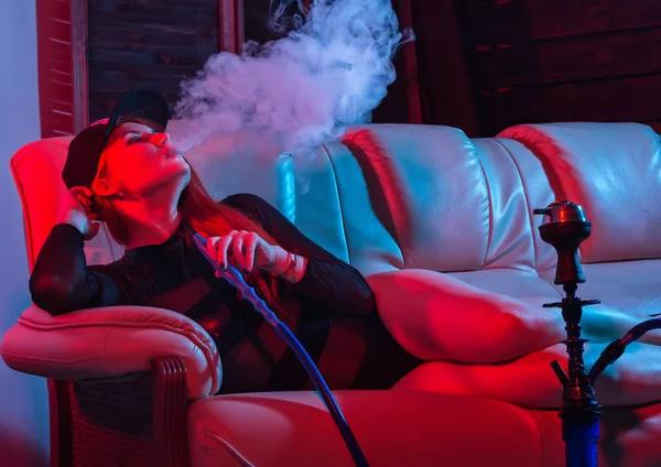 Sexy zrzavá žena v černém obleku sedí na pohovce a kouří Hookah, holka vyžene velký kouř, červené modré tóny — Stock fotografie
