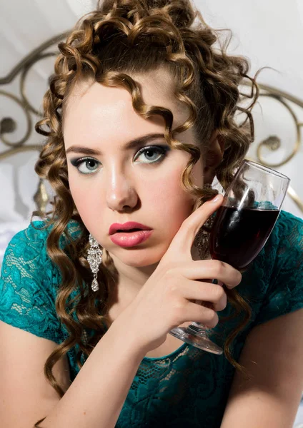 mysterious young curly woman resting on the bed and drinks red wine