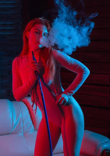 Sexy redhead hipster woman vaping electronic hookah. girl blows out big smoke in a red blue tones. — Stock Photo, Image