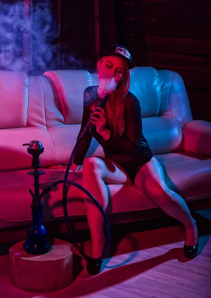 Sexy rothaarige Hipsterfrau im schwarzen Body sitzt auf einem Sofa und raucht Wasserpfeife, Mädchen bläst dicken Rauch in einem rotblauen Farbton aus — Stockfoto