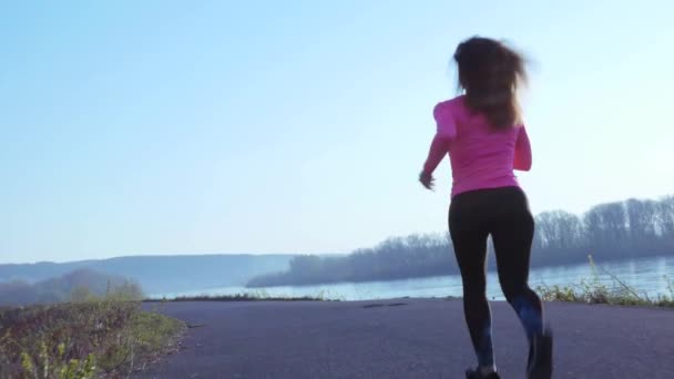 Sportive girl jogging along the river bank during sunrise or sunset. healthy lifestyle concept of athletic woman goes in for sports. slow motion — Stock Video