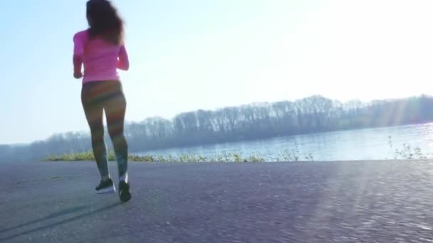 Sportive girl jogging along the river bank during sunrise or sunset. healthy lifestyle concept of athletic woman goes in for sports. slow motion — Stock Video