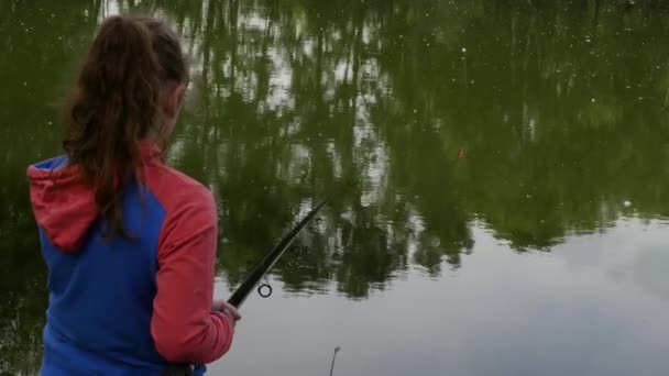 Roztomilá mladiststná dívka lovící na břehu řeky stojící na písku a držící rybářské tyčinky. — Stock video