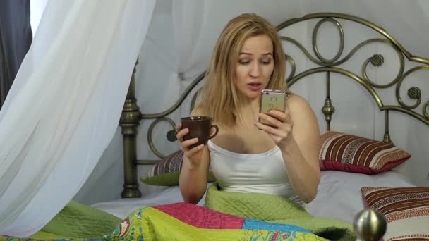 Veselá žena sedí v posteli tancovat a zpívat, pití kávy a chatování s přáteli na telefonu. 4k — Stock video