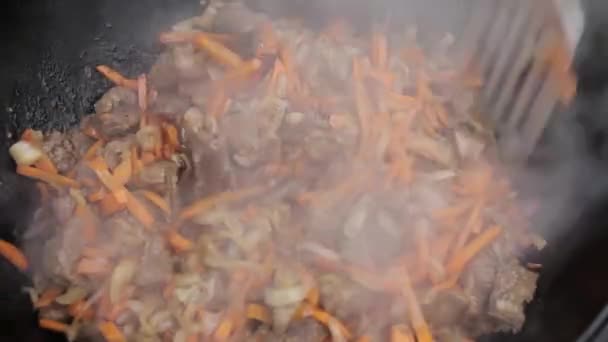 Cozinhar de pilaff com cordeiro, cenouras e temperos cebolas em um caldeirão em um fogão a lenha — Vídeo de Stock