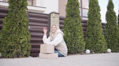 Uzun yeşilliklerle çevrili modern bir evin yanında kutularla oturan teslimatçı kadın.