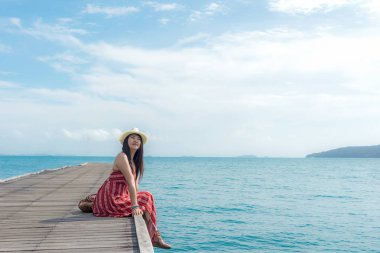 Yaz günü. Kadın gülümsüyor rahatla ve kırmızı giyen elbise moda ahşap köprü'de deniz, mavi gökyüzü arka plan üzerinde oturuyor. Seyahat ve tatil. Özgürlük kavramı