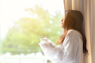 Asyalı kadın çevre pencere, uyan sonra kahve içme yatak içinde güneşli sabah. Yaşam tarzı kavramı.