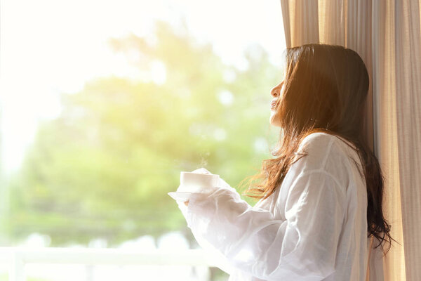 Asian woman in bedroom drinking coffee after wake up near window, sunny morning.  Lifestyle Concept.