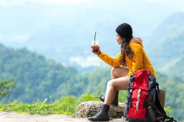 亚洲女性徒步旅行者或带着背包探险的旅行者坐在那里 喝着冰镇咖啡 放松下来 在山上休息 到户外度假 享受目的地的休闲教育 旅行和生活方式概念 — 图库照片