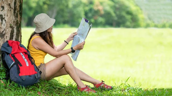 Asian women hiker or traveler with backpack adventure holding map to find directions and sitting relax in the jungle forest outdoor for education nature on vacation. Travel and Lifestyle Concept