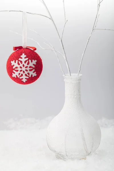 Red ball with the image of a snowflake on a white branch in a frost-covered white vase on a background of white snow