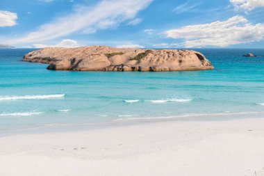 Stone island in the azure waters of the ocean against a blue sky with white clouds and a sandy beach in the foreground clipart