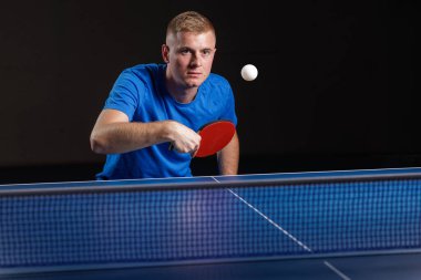 Mavi tişörtlü erkek sporcu, kapalı masa tenisi maçında kırmızı kürekli topu geri getiriyor. Rekabet ve antrenörlük temaları için ideal.