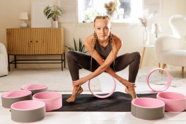 Kadın, omurga hareketlilik dengesini ve iyileşme sürecini geliştirmek için evdeki derin bir çömelmede yoga tekeriyle ısınıyor.