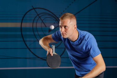 Genç adam masa tenisi servisine hazırlanıyor, top kapalı bir kulüpte havada süzülüyor. Spor reklamları ve koçluk içeriği için dinamik eylem ve kopyalama alanı.