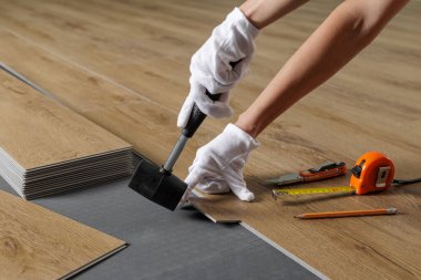 Akustik alt katmandaki SPC plastiğini döşeme çalışmaları sırasında kauçuk çekiç ve aletlerle hizalayan ellerin yakın görüntüsü