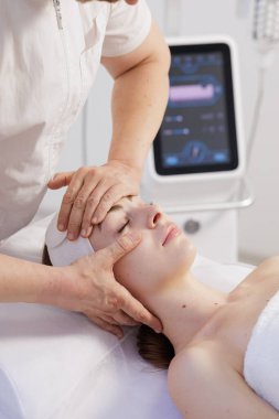 Kadın estetisyen ve cihaz geçmişi olan temiz bir klinikte yüz masajı sırasında rahatlıyor. Sağlık ve cilt bakımı projeleri için ideal.