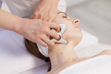 Profesyonel estetisyen, klinik tedavisini gevşetmek için paslanmaz gua sha kazıyıcı ve taze parlatıcı ile yüzünü yontuyor.