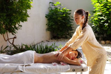 Terapist bir spa bahçesinde sırt masajı yapıyor, doğal ışık ve orkideler sağlığını, sakinliğini ve konukseverliğini iletiyor..