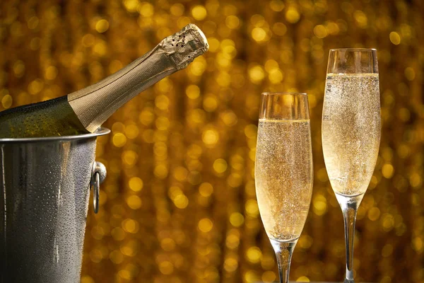 Bottiglia piena di champagne in un secchio con ghiaccio e due bicchieri di champagne su sfondo bokeh dorato — Foto Stock