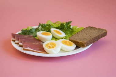 Kahvaltı. Haşlanmış yumurta, pastırma, pembe bir arka plan üzerinde beyaz bir tabak içinde Yeşiller.