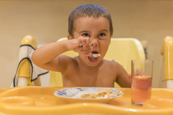 这个男孩2年吃粥 儿童餐桌 儿童独立的概念 可爱的小男孩用勺子是酸奶 孩子笑了 婴儿座椅上的滑稽孩子 — 图库照片
