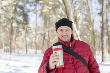 Adam fincandan çay içiyor. Kış ormanı Gri hırkalı, örgü şapkalı ve eşarplı genç adam karlı arka planda duruyor ve elinde çay fincanı var.