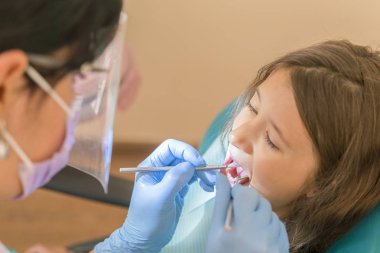 Bir dişçi koltuğu yalan ve bir sonda kullanarak bir diş hekimi tarafından yürütülen bir ağız boşluğu muayenesi yaparken kameraya gülümseyen sevimli küçük kız. Küçük kız dişçi ofisinde oturuyor.