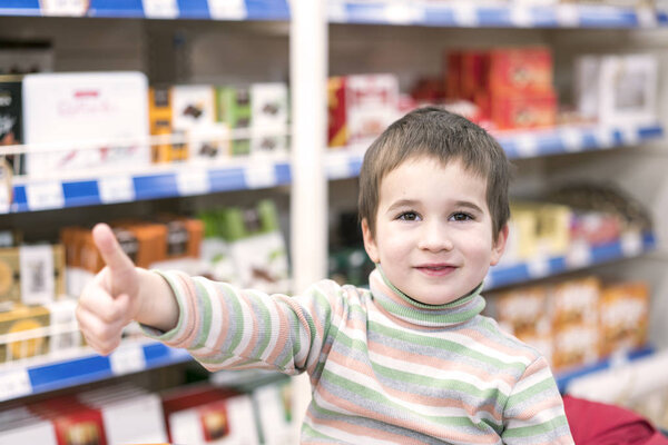 Happy boy 4 years in a supermarket on the background of shelves with chocolate. The boy showed that everything is ok.