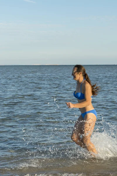 Μια σέξι μελαχρινή νεαρή γυναίκα ή κορίτσι που φοράει ένα μπικίνι που τρέχει μέσω του surf σε μια έρημη τροπικό παραλία με ένα γαλάζιο ουρανό. Νεαρή γυναίκα τρέχει δίπλα στη θάλασσα. κάθετη φωτογραφία — Φωτογραφία Αρχείου