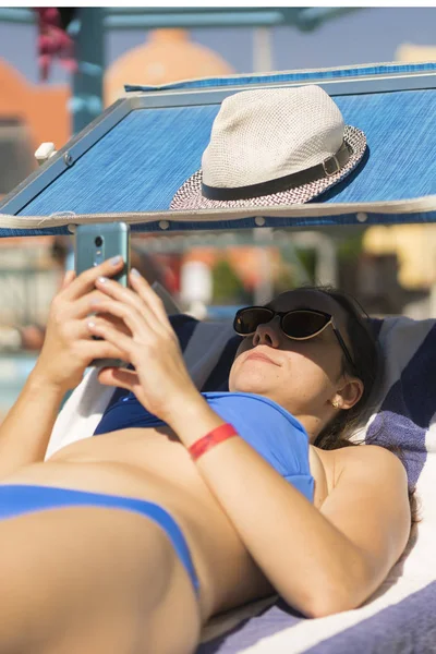 Sommer-Lifestyle-Porträt einer jungen atemberaubenden Frau, die auf der Sonnenliege am Strand der tropischen Insel liegt und ihr Smartphone benutzt. junge weiße Frau auf blauer Sonnenliege liegend — Stockfoto