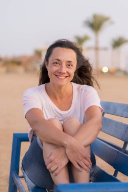 Bankta oturan genç kadın. Mavi bir Bank üzerinde güzel bir gülümseme olan bir kadın. Huzur ve mutluluk kavramı. dikey fotoğraf